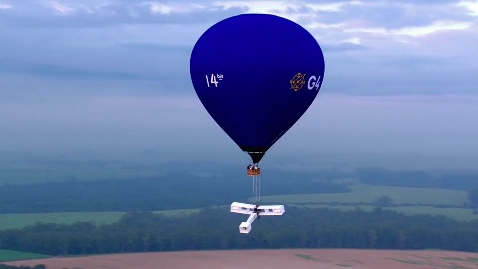 Balão azul carrega a réplica do 14-bis sobre um campo verde visto do alto